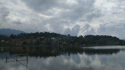 A wide, tranquil landscape shot captures a lake or large body of water in Kecamatan Panjalu, Indonesia, under a heavily overcast sky. The foreground is dominated by the calm, reflective surface of the water, mirroring the grey clouds above and the distant landmass. To the left in the water, a simple, partially submerged bamboo structure, possibly a fishing platform or part of a fish farming setup, stands out.

In the midground, a village is nestled along the water's edge, surrounded by dense, lush green trees and foliage. Several houses with distinct reddish-brown tiled roofs are visible among the greenery, indicating a populated area. Near the village, along the shore, some structures are visible in the water, suggesting aquaculture facilities like fish cages.

The background features a range of mountains or large hills on the left side of the frame, their peaks gently rising and partially obscured by the low, thick clouds. The entire landscape, from the village slopes to the distant mountains, is covered in rich, verdant vegetation. The sky is completely blanketed by voluminous grey clouds, contributing to a soft, diffused lighting and a serene, albeit muted, atmosphere typical of an overcast day. There is no visible human activity or text in the image.