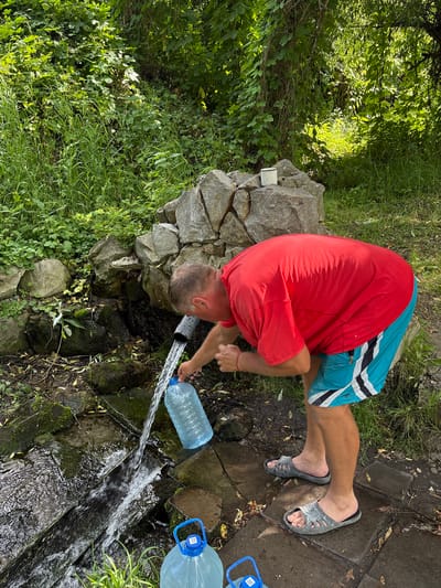 In Новоподклетное, Russia, a man wearing a red t-shirt and blue and white striped shorts is filling a large plastic water bottle with water from a natural spring. The spring is located near a rock formation and is surrounded by lush green foliage. Two other large water bottles are visible in the foreground. The man is using a gray hose to direct the water into the bottle. The scene appears to be taking place outdoors in a natural setting.