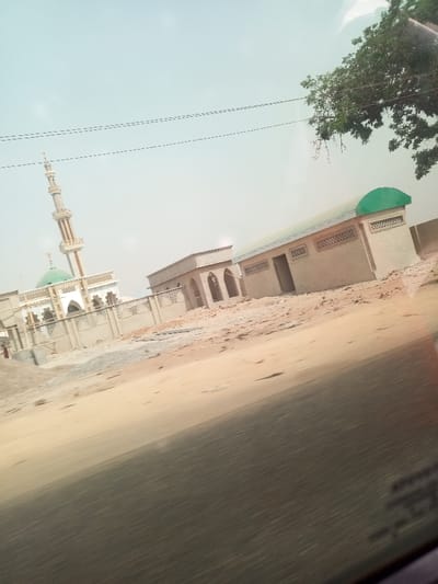 The image captures a daylight scene, likely in Uki, Nigeria, focusing on architectural structures in a dry, open environment. The primary subject is a mosque, prominently featuring a tall, slender minaret with a crescent moon finial and a green dome. The mosque's main building is adorned with arched gateways and a decorative perimeter wall, appearing newly built or under construction.

Adjacent to the mosque, to the right, are several single-story buildings constructed from concrete blocks. These structures also suggest recent construction or development, exhibiting arched entrances and patterned vents. One of these buildings features a green roof or awning, complementing the mosque's dome. The ground across the foreground and surrounding the buildings is composed of exposed sand and dirt, indicating a dusty or barren landscape.

The sky is bright and appears light blue or hazy white. Black power lines are visible stretching across the upper part of the sky, and a portion of a green-leaved tree branch is seen in the upper right corner. The blurred, dark area in the immediate foreground suggests the photograph was taken from inside a moving vehicle. No people are visible in the scene, and there is no discernible text on any of the buildings or structures. The overall impression is one of ongoing development and community infrastructure in a dry, possibly rural or semi-urban setting.