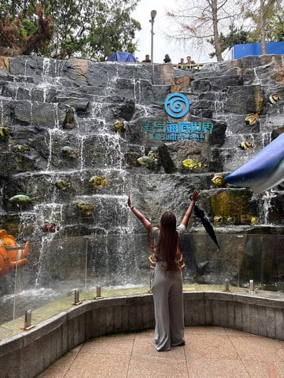 The image depicts a female individual standing with her back to the camera, arms raised in a "V" shape, facing a large artificial rock waterfall at the Qingdao Underwater World in Shinan District, Qingdao, China. She has long, reddish-brown braided hair and is wearing a light grey, loose-fitting two-piece outfit consisting of a crop top and wide-leg pants. A closed black umbrella is held in her left hand.
The multi-tiered waterfall cascades into a pool behind a curved, low concrete barrier topped with a transparent glass panel supported by metal stanchions. Embedded into the dark grey rock face of the waterfall are numerous colorful artificial fish models of various species. A prominent blue spiral logo and the white text "青島海底世界" (Qingdao Underwater World) are visible on the rock face above the waterfall's midpoint. At the lower left, a large orange clownfish model is partially visible, while a large blue and white shark head model is positioned on the lower right, extending into the frame.
Above the waterfall, on a raised platform or walkway, several indistinct individuals are visible, along with a blue tarp. The area behind and above the waterfall features green trees and some deciduous trees with bare branches, suggesting an outdoor environment. An outdoor lamppost is visible on the upper left. The sky is overcast and grey. The ground in the foreground is a light-colored tiled surface.