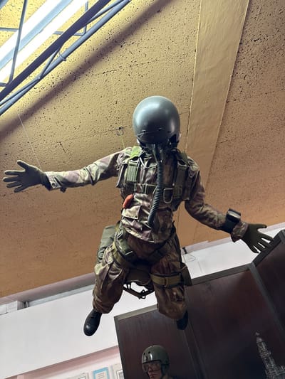 The image displays a military-dressed mannequin suspended in an indoor environment in Praia do Ribatejo, Portugal. The central subject is a mannequin outfitted in a camouflage flight suit, a dark green pilot's helmet, and an oxygen mask with a ribbed hose connected to the face. Dark gloves cover both hands, which are outstretched laterally. A harness system with a backpack-style parachute pack and an orange cylindrical object is affixed to the suit. A black device is secured to the mannequin's left wrist. Its legs are bent at the knees, and it wears dark boots. The background features a textured, light brown ceiling with visible fluorescent light fixtures and dark overhead structural beams. Below the suspended mannequin, a dark wooden display cabinet occupies the right foreground, behind which a second mannequin's head, wearing a green helmet and clear goggles, is partially visible. A white wall with framed items is visible in the lower-left background.
