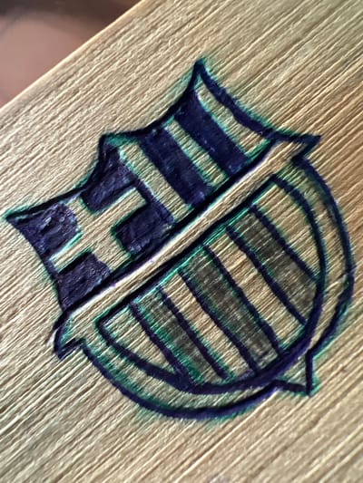 The image displays a close-up, angled shot of a shield-like emblem with vertical stripes. The emblem is rendered in dark blue and green ink on a textured, light-colored surface, possibly wood or paper. The lines of the emblem are thick and somewhat uneven, suggesting it might be hand-drawn or stamped. The texture of the underlying surface is visible, with fine, parallel lines running diagonally across the image. The emblem appears to be divided into two sections, with the left side featuring a cross-like shape and the right side showing a series of parallel lines within a curved border. The lighting is somewhat dim, creating shadows that emphasize the texture of the surface and the depth of the ink lines. The photograph is focused on the emblem, with the background blurred. There are no people, actions, or specific environmental features like buildings or landmarks visible in the image. This image is a detail of an emblem and does not provide any specific information about its location in Villa de Cura, Venezuela, or any other geographical context.