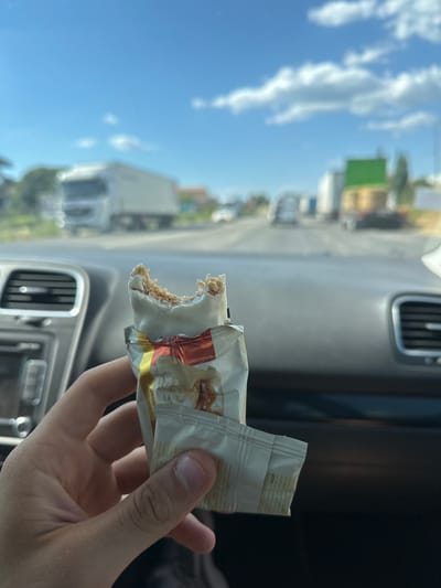 A hand is holding a partially eaten nutrition bar, its white chocolate coating broken to reveal a granola-like interior. The bar is in its foil wrapper, which has some text and graphics, including the words "Illustrative Image". The background suggests the setting is inside a vehicle, as indicated by a car's dashboard with air vents and a steering wheel. Through the vehicle's windshield, a bright, sunny day is visible. Fluffy white clouds dot a clear blue sky. The landscape outside appears to be a semi-urban or roadside environment, with buildings, trees, and a large truck in the distance. The lighting suggests it's daytime. The image is oriented sideways.