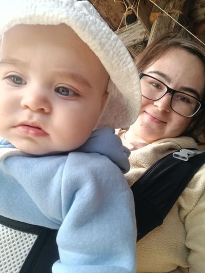 A close-up, eye-level shot depicts a young woman holding a baby. The woman has light skin, brown hair, and wears glasses. Her face is turned slightly towards the camera, and she is smiling faintly. She is wearing a tan-colored zip-up sweatshirt over a pinkish-beige t-shirt. A black baby carrier strap is visible across her chest. The baby, who is the primary focus of the image, is positioned in the carrier. The baby wears a light blue hooded sweatshirt and a white bonnet-style hat. The baby's face is turned towards the camera, and the baby has a serious expression.
The background includes wooden structures and some dried foliage.
There is no verifiable indication that the location of the image is in the city of Усть-Тунтор, Russia.