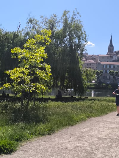 The image depicts a park-like setting in Chézenas, France. In the background, buildings are visible, including a church with a tall spire. A tree with bright green leaves stands prominently in the foreground, with weeping willow trees behind it. A body of water, likely a river or a large pond, is also visible, with people engaging in water activities. A dirt path runs along the edge of the park, and a person is walking along it. Grass and other greenery cover the ground. People are seated along the waterfront. The sky is blue with some clouds.