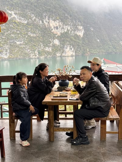 An outdoor scene in Gongtan, China, features four individuals seated at a wooden table on a tiled patio overlooking a body of water and heavily forested mountains under an overcast sky.

From left to right:
A young female child, wearing a black puffer jacket, dark blue pants, and light-colored sneakers, sits on a wooden chair, facing right with hands resting in her lap.
Adjacent to her, an adult female with long dark hair tied back, dressed in a dark long-sleeved garment, sits on a wooden bench, smiling and looking towards the man across the table. Her hands are positioned on the table surface.
An adult male with short dark hair, wearing a black leather jacket, dark pants, and black sneakers, sits on a wooden bench, holding a small transparent cup to his mouth and looking directly at the camera.
Partially obscured behind the man, an adult female with long dark hair covered by a light-colored baseball cap, wearing a dark long-sleeved top, sits on the wooden bench, looking towards the man and smiling.

The rustic wooden table holds a dark ceramic vase containing decorative dried branches, multiple small ceramic tea cups, a glass teapot filled with amber liquid, a wooden tray containing small, light-colored snacks, and a dark mobile phone. The patio features a dark tiled floor and a wooden railing structure. A red Chinese lantern is visible suspended from the upper left.

The background reveals a serene body of water, likely a river or lake, reflecting the grey sky. This water is bordered by steep, rugged mountains densely covered in green vegetation, with some visible rock faces. The sky above is uniformly overcast and hazy. On the far right, a complex of structures with red roofs is visible on the waterline.