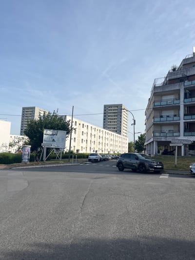 The image depicts a street scene in the city of L'Haÿ-les-Roses, France, likely in a residential area with high-rise apartment buildings and lower-rise apartment blocks. In the foreground, a paved intersection is visible with a pedestrian crossing. A dark grey Citroën C5 Aircross SUV is parked on the right side of the road, partially on the pedestrian crossing, with its license plate visible as NJ 956 SL. To the left, a white vehicle is partially visible. In the middle ground, a man in dark shorts and a t-shirt is walking on the sidewalk. Behind him, a large billboard displaying architectural renderings and text is mounted on a metal frame. A tall, slender streetlamp stands to the right of the billboard. Further back, several multi-story apartment buildings are present, with one tall, light-colored building dominating the skyline in the distance. The sky is a clear, pale blue with wispy clouds. A signpost pointing to the right indicates "Centre Socio Culturel Eglise St Paul" and "AVARIA."