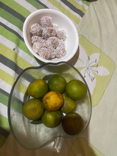 A close-up, top-down shot shows a bowl of small, round candies coated in shredded coconut next to a clear glass bowl filled with tangerines on a striped fabric surface. The candies, appearing to be about an inch in diameter, are clustered in a white bowl. The tangerines, varying in color from green to yellow and some with browned spots, are arranged in the clear bowl, which has a decorative, wavy rim. The fabric beneath has a pattern of green, white, and gray stripes on the left, transitioning to a lighter green fabric with a stylized white flower design on the right. The image is taken from an overhead perspective. This scene is located in Anggaswangi, Indonesia.