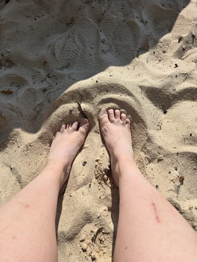 A close-up, overhead perspective shows a person's bare feet and lower legs partially submerged in light-colored sand. The sand shows ripples and indentations, including a distinct shoe print in the upper left quadrant of the image. A shadow, likely from an overhead object or structure, falls across the sand in the upper half of the frame, obscuring some of the detail. The skin tone of the legs is fair, and a faint purplish mark, possibly a scar or bruise, is visible on the right leg. The overall environment suggests an outdoor setting, possibly a beach or sandy park area.