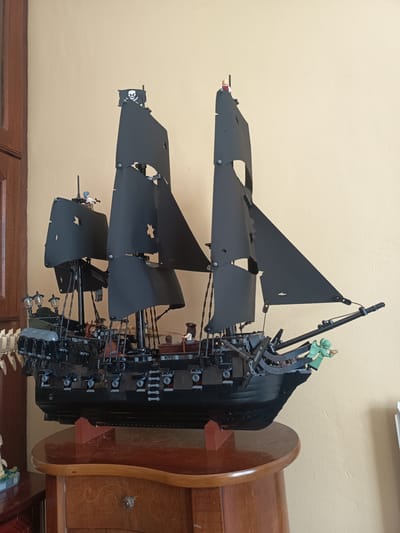 A Lego model of the Black Pearl pirate ship is displayed on a wooden table in a room. The ship is predominantly black with black sails, some of which are tattered. A Jolly Roger flag with a skull and crossbones is visible at the top of the main mast. Several Lego minifigures are present on the deck of the ship, including what appears to be a figure in a red jacket and a figure in green at the bow. The ship is supported by two brown Lego brick stands. The background consists of a plain, light-colored wall and a wooden door on the left side of the image. The scene appears to be an indoor setting, possibly a display in a home or a collector's space. The image does not provide any information or context to determine its location in the city of Millstreet, Ireland.