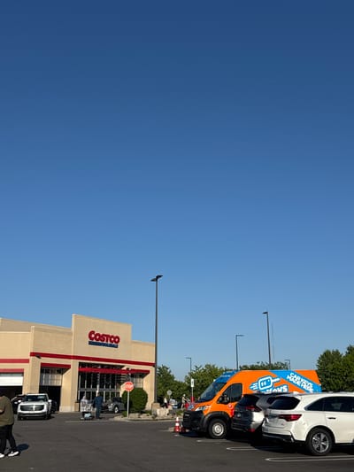 The image shows the Costco Wholesale store in Bloomingdale, Illinois, USA. The Costco building is beige with red trim around the entrance. Customers are visible near the entrance, some with shopping carts. In the parking lot, vehicles are parked, including an orange van with "Car Keys Made Here" printed on the side. The sky is clear and blue. Streetlights are visible. The image is taken during the day.
