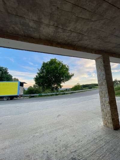 A wide-angle, eye-level outdoor shot captures a serene landscape with a focus on a road and a truck, framed by a concrete overhang and a stone pillar on the right. The scene appears to be located on the outskirts of Valkosel, Bulgaria.
In the foreground, a rough, concrete surface extends towards the viewer, showing some texture and scattered dark spots. To the right, a thick, square pillar constructed from light-colored, rough-hewn stones stands prominently, supporting the overhead structure. The overhang above is made of gray concrete, with visible lines and imperfections.
Beyond the foreground, a paved road curves gently through a verdant landscape. On the left side of the road, a truck with a bright yellow cargo box and a blue trim is visible, moving away from the viewer and towards the right. The truck is partially obscured by its motion blur.
The middle ground features a cluster of lush green trees, with one particularly large, mature tree with a full canopy dominating the center of the frame. A silver guardrail runs along the side of the road, separating it from the rolling hills in the background.
The sky above is a picturesque blend of soft blue with scattered white and wispy clouds, tinged with the warm hues of either sunrise or sunset. The overall atmosphere is tranquil and natural, suggesting a rural or semi-rural setting. The presence of the truck and the unfinished concrete structure hint at ongoing development or activity in the area. There are no people visible in the image.