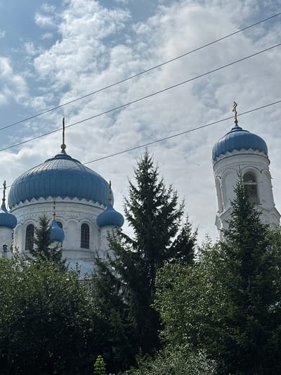 The image shows the bell tower and main dome of an Orthodox church, likely located in the city of Nogornyi, Russia. The church features white walls and bright blue domes topped with golden crosses. Surrounding the church are lush green trees, including pine trees. The sky is cloudy, with patches of blue visible. Several power lines stretch across the upper portion of the image. No people or specific actions are depicted.