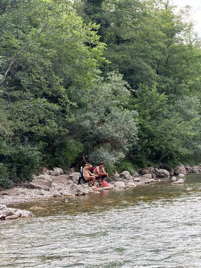 Two people are sitting on chairs on the rocky bank of a river. The person on the left is wearing black shorts and a tan shirt, and is holding a yellow object. The person on the right is wearing black shorts and a red top. There are pink objects near the person on the right. The river is wide and appears to be flowing quickly, with light reflecting off its surface. Behind the people is a dense area of trees and vegetation, with some larger rocks interspersed. The scene appears to be set during the daytime, under what looks like overcast skies. The overall impression is of a relaxed, outdoor setting.