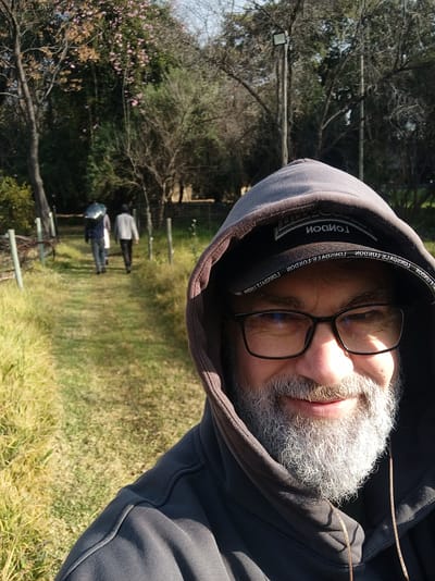 The media file is a selfie taken by a person wearing a dark grey hooded jacket and glasses. The person has a grey beard and appears to be looking directly at the camera. The background is a grassy area with trees and two other people walking in the distance. There is also a light fixture visible on a pole in the background. The scene appears to be outdoors during the daytime, with sunlight casting shadows. The overall mood seems casual and relaxed. Text is visible on the inside of the hood, reading "LONDON". The location context is Randburg, South Africa.