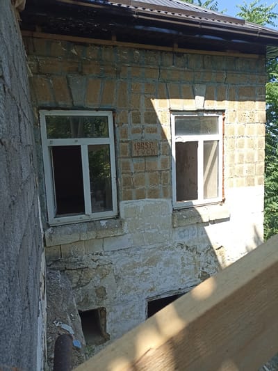 This media file captures an exterior view of a weathered, stone building with two open windows on its upper level. The building appears to be constructed from light-colored, porous stone blocks, some of which are chipped or cracked, suggesting age or disrepair. The lower portion of the building is covered in peeling white plaster. To the left, a rough, grey concrete wall is visible, hinting at an adjacent structure or an unfinished section. Below the windows, a dark, rectangular opening is present in the wall, possibly a small cellar or utility access.

In the center of the stone wall, between the two windows, a raised inscription is visible. It appears to read "1965 r. V.G.M.". The inscription is weathered and partially obscured by shadows.

The scene suggests a rural or semi-abandoned setting, with trees and foliage visible in the background through the windows and to the right of the building. The lighting indicates it is daytime, with strong sunlight casting distinct shadows on the building's facade. The overall impression is one of an old, possibly historical structure in need of maintenance, set in a natural environment. The presence of a wooden plank or beam in the foreground suggests ongoing construction or renovation work, or perhaps debris from a past project. No people are visible in the image.