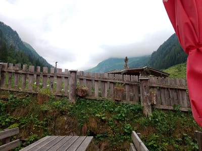 The image shows a weathered wooden fence in the foreground, with lush green vegetation growing on the bank behind it. Beyond the fence, a rustic wooden structure with a roof is visible, and further in the background, a mountain range covered in dark green pine trees stretches into the distance, partially obscured by clouds. A bright red fabric, possibly an umbrella or awning, is visible in the upper right corner, partially obscuring the view of the mountains. The overall scene suggests an outdoor setting in a mountainous region, likely a rural or alpine location. While the image does not explicitly show Bramberg am Wildkogel, Austria, the visual elements are consistent with the landscape of the Austrian Alps, a region where Bramberg am Wildkogel is located.