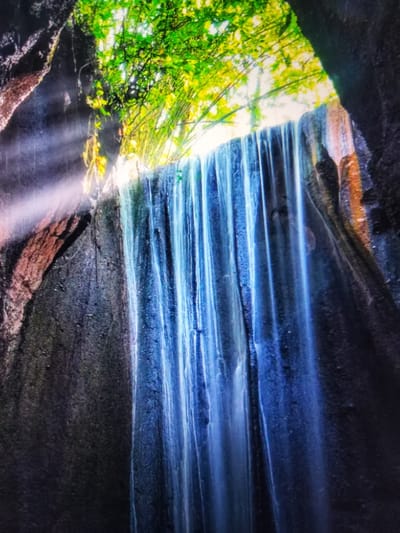 The image shows a waterfall cascading down a rocky cliff face. The water is a vibrant blue, and light rays are visible filtering through the trees above. The rocks are dark and textured, with some patches of reddish-brown. The foliage at the top is lush and green, with some yellow highlights. The scene appears to be a natural setting, possibly a cave or grotto, with sunlight breaking through an opening. The description of "Payagaboh, Indonesia" in the prompt does not correspond to any known location and is therefore not included in the description of the image.