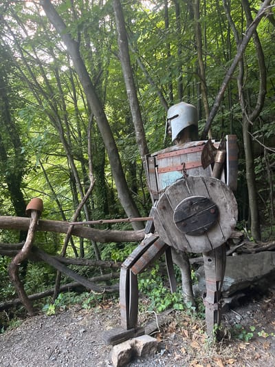The image depicts an outdoor scene, likely in a wooded area, with the camera oriented vertically. The primary subject is a rustic, life-sized effigy or sculpture constructed from reclaimed wood and metal. It appears to be a humanoid figure, possibly an automaton or a scarecrow, with a cylindrical body made of wooden staves reinforced with metal bands. A large wooden cable spool serves as a prominent feature, possibly representing a torso or a shield. The figure has a headpiece resembling a helmet, made of a curved piece of metal. Attached to the effigy is a metal chain, and what looks like a small, worn metal tag or plaque. A rustic tool, possibly an axe head, is also affixed to the structure. The effigy is surrounded by trees and vegetation, with a gravel path visible in the lower right portion of the image. The setting suggests a rural or natural environment, possibly a garden, yard, or park area in Golyamo Belovo, Bulgaria. The lighting suggests it is daytime, with dappled sunlight filtering through the tree canopy. The overall impression is one of an artistic or decorative installation, perhaps with a historical or folk theme, given the materials used.