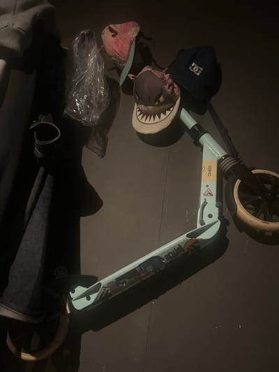 The image depicts a scooter lying on the ground in a dimly lit environment. Several objects are scattered near the scooter, including a dark blue cap with a "DC" logo, a hat with shark teeth design, a pink cap, and a clear plastic bag. The scooter is light blue and has stickers on its frame. The location is Cangkuang Kulon, Indonesia. Other items include a jacket and what appears to be a bag.