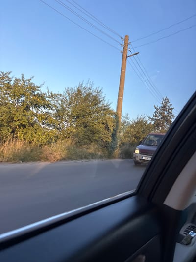 The image depicts a view from inside a vehicle, looking out at a rural road in Slaveevo, Bulgaria. The scene is characterized by a clear blue sky, suggesting fair weather, and possibly late afternoon or early evening given the warm light.

The primary objects in the scene are a wooden utility pole with wires stretching across it, and a maroon van with a visible license plate. The van appears to be traveling on the paved road, which runs alongside a dense line of trees and vegetation. The trees are lush and green, with some hints of yellow, suggesting late summer or early autumn. There are also some taller trees in the background. The foreground of the image is dominated by the interior of the car, including the window frame and a portion of the dashboard, indicating the perspective of someone being driven.

The license plate on the van clearly displays the text "TX 0837". The image captures a moment of transit, with the van moving along the road. The overall impression is one of peaceful, everyday travel through a bucolic landscape. There are no discernible people in the image apart from what appears to be a silhouette of someone in the driver's seat of the van.