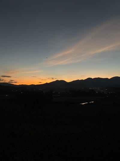 The scene depicts a twilight landscape in Ban Tung Yao, Thailand, during sunset. The sky exhibits a gradient from dark blue-grey overhead to orange and yellow at the horizon, with two prominent, elongated, light-colored clouds extending diagonally across the upper half. A small, dark, indistinct flying object is visible against the sky, slightly left of center. A silhouette of undulating mountains forms the dark horizon. Below the mountains, scattered points of artificial light delineate a village settlement in the midground, with faint wisps of smoke or haze visible near some lights. A narrow, reflective body of water is present in the lower mid-right. The foreground consists of dark, undifferentiated land or fields, devoid of specific detail. No individuals or direct actions are depicted.