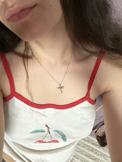 This media file shows a close-up, overhead shot of a young person lying down, with their head tilted to the side. The person has long, dark hair and is wearing a white tank top with red trim. A delicate silver necklace with a small cross pendant rests on their collarbone. The tank top features a graphic of two red cherries with green leaves on a branch.
The setting appears to be indoors, likely a bedroom, as indicated by the presence of bedding with a textured, ribbed pattern. The lighting suggests it could be daytime, with natural light illuminating the scene. The overall mood is relaxed and casual. There is no visible text in the image. The location context is Postoyata, Ukraine.