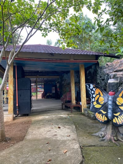 The image shows a view from the entrance of a building in Kecamatan Manyaran, Indonesia, with a large painted butterfly sculpture in the foreground. The building is made of concrete and has a tiled roof. The entrance is covered by a roof overhang, supported by pillars. The building is decorated with a blue and white mural on the interior wall. The concrete walkway leading to the entrance is paved with grey concrete. The sculpture is made of painted concrete, with black, yellow, and red accents. The sculpture is mounted on a concrete base. The background is a green, leafy, tropical landscape with a large tree dominating the left side of the image. The building appears to be a commercial building with a small, open-air dining area. There are no people in the image.