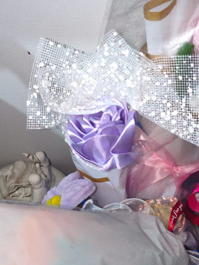 This media file captures a close-up view of a gift, likely a bouquet or decorative arrangement, with a soft, pastel color palette dominated by purples, pinks, and whites. The central focus is a delicate lavender rose, crafted from satin fabric, nestled within a white net fabric adorned with shimmering silver glitter. To the left, a portion of a light-colored ceramic figurine, possibly an angel, is visible. Further to the right, a pink ribbon is tied in a bow. A small red ribbon with white text that reads "just for you" is also visible, adding a personal touch. The overall aesthetic suggests a thoughtful and celebratory gift. The background is indistinct, with soft lighting that creates a gentle, almost ethereal mood. No specific location cues are apparent beyond the context of a gift, but given the provided location context of Cavite City, Philippines, this gift could be for any occasion celebrated there. The time of day and weather are not discernible.