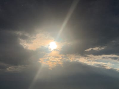 The image depicts a dramatic sky with the sun partially obscured by dark, heavy clouds. A bright beam of sunlight pierces through the clouds, creating a luminous ray that extends diagonally across the frame. The clouds themselves vary in darkness, with some areas appearing almost black, while others are illuminated by the sun's rays, taking on a yellowish or golden hue. The overall impression is one of a dynamic and possibly stormy sky. The location is specified as Somoniyon, Tajikistan. There are no people, specific subjects, objects, or actions visible in the image; it is solely a view of the sky.
