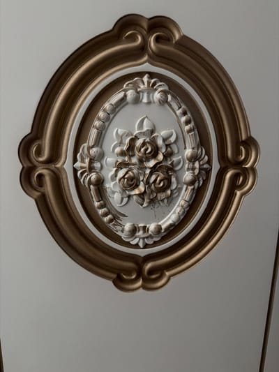 This media file is a close-up, eye-level shot of an ornate oval-shaped decorative element, likely a ceiling medallion, with a matte white background. The medallion is framed by a thick, metallic gold molding with a subtle sheen, featuring a stylized, flowing design. Inside the gold frame, there's a recessed oval area with a white, textured surface. At the center of this inner oval, a cluster of three rose-like floral decorations stands out. These roses are rendered in a metallic gold finish, contrasting with the white background and the outer gold frame. The roses appear detailed, with carved petals, and are interspersed with smaller decorative elements that mimic leaves and buds, also in gold. Some areas of the white surface show faint smudges or imperfections, suggesting age or a patina. The lighting is soft and diffused, casting gentle shadows that highlight the relief and texture of the medallion. There is no visible text. The overall impression is one of decorative artistry, likely found in an interior setting. The metallic accents and floral motifs suggest a classical or baroque aesthetic.