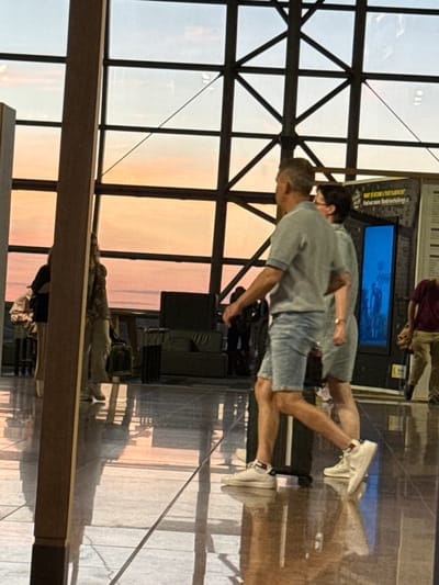 The image depicts a group of people in an indoor environment with large windows that reveal a sunset or sunrise sky with orange and blue hues. The architecture suggests a modern public space, possibly an airport or a large hall, with a tiled floor that reflects the overhead lighting and the colors from the sky.
In the foreground, a man and a woman are walking side by side. The man is wearing a light gray striped polo shirt and light blue denim shorts, and white sneakers with red and blue accents on the socks. He is pulling a black suitcase. The woman, to his right, is wearing a light gray dress and white sneakers. They appear to be walking away from the viewer.
Further back, to the left, another person, possibly a woman, is standing with a bag slung over her shoulder. She is wearing a patterned top and light-colored pants. Behind her, a dark armchair or sofa is visible.
To the right, a person in a dark red shirt and khaki pants is walking away from the viewer. There are also displays or posters on the wall in the background, one of which has a bright blue illuminated section. The overall scene is brightly lit, with strong reflections on the floor.
The location is identified as Zaventem, Belgium. Given the presence of luggage and the large windows suggesting a travel hub, this scene is likely within Brussels Airport, which is located in Zaventem, Belgium.