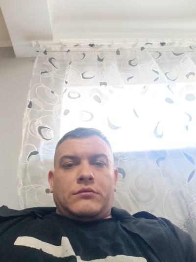 The image shows a selfie taken indoors, likely in a residential setting. A man with short, dark hair and a light complexion is the primary subject, positioned centrally in the frame and looking directly at the camera. He is wearing a black garment, possibly a t-shirt or jacket, with a white graphic design on the front.
The background consists of a white ceiling with a slight slant, a curtain rod, and sheer white curtains with a repeating circular, abstract pattern in black and grey. A window is visible behind the curtains, letting in bright light that illuminates the scene. The overall lighting is somewhat harsh due to the bright light source behind the subject.
The location is identified as посёлок совхоза "Буденновец", Russia. This suggests the image was taken in or around this specific settlement. The indoor environment, with its typical domestic furnishings like curtains, implies a personal space such as a home. There are no other individuals or animals present in the image. The composition is a close-up of the man's upper body and face, with the background serving as context.