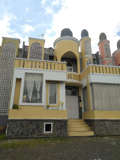 The image displays the exterior of a multi-story building exhibiting an eclectic architectural style, primarily painted yellow with white trim and dark gray accents, under a partially cloudy sky. The foundation of the building is clad in rough, dark gray stone veneer. A small, dark, rectangular window with a white frame is visible in the lower left section of the stone foundation.
The ground floor, painted yellow, features a large rectangular window with white framing and internal sheer white curtains on the left. A smaller, vertical rectangular window is positioned next to it. The central entrance consists of five yellow-painted steps leading to a light gray arched door with a semicircular transom window above. Two thin, light-colored plants stand on the yellow wall to the right of the door. Another large rectangular window with white framing and light-colored sheer curtains is present on the right section of the ground floor.
Above the ground floor, a yellow balcony railing with vertical balusters spans the width of the structure. Behind this railing, to the left, is a section of wall adorned with intricate dark gray and white patterned screen panels. The central and right portions of the upper level are characterized by rounded yellow structures, one featuring an arched alcove. These are topped by cylindrical towers with dark gray conical roofs. Multiple similar cylindrical towers, some with patterned screen panels and one distinctly peach/pink in color, extend across the roofline to the right. A television antenna is visible on the roofline above the left patterned wall section.
The foreground consists of dark gray paved ground with sparse green grass and weeds. No individuals, actions, or specific interactions are visible in the scene. The image does not contain any discernible information to confirm its location in the city of Nathene, Laos.