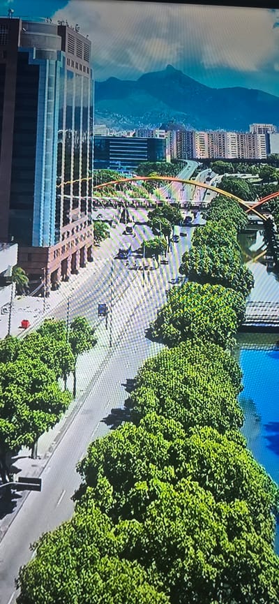 The image depicts an aerial view of a city scene, featuring a tall, modern skyscraper on the left with a patterned facade. In the background, mountains are visible under a cloudy sky. The foreground shows a road with several cars driving on it, lined by lush green trees. A body of water, appearing blue, is situated to the right of the trees. There is an orange bridge spanning across the road and water, also lined with trees. The scene is a daytime outdoor environment. There is no indication that this image is related to Lippstadt, Germany.