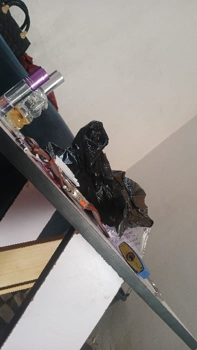 A cluttered shelf displays a variety of items. There's a black bag, assorted cosmetics including perfume bottles, a brown belt with a buckle, and what appears to be a small lock with a blue handle. These items are arranged haphazardly on the shelf, suggesting a lack of organization. The shelf is positioned at an angle, creating a dynamic composition.