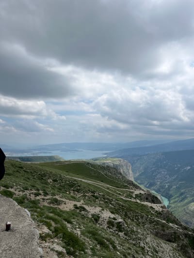 The image depicts a scenic view from a high vantage point, possibly a mountain or cliffside in Chirkey, Russia. The sky is overcast with prominent clouds. A body of water, possibly a river or reservoir, snakes through the landscape below. The terrain consists of steep, mountainous slopes covered with patches of green vegetation and bare rock. Faint trails or paths are visible along the hillsides. The foreground features a concrete structure, likely a part of the observation point.