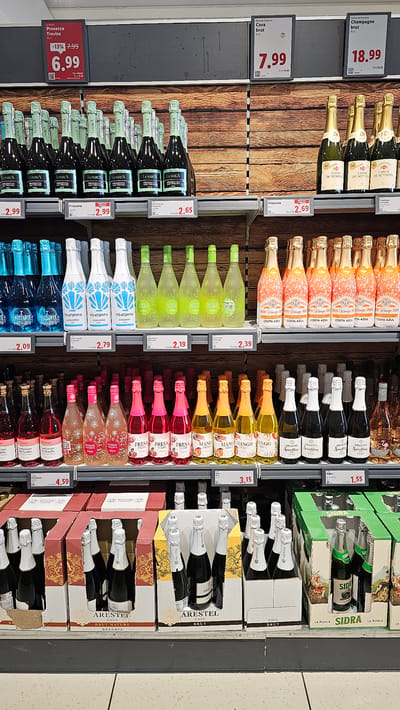The image shows a shelf display of alcoholic beverages in a supermarket, likely in Cuarte de Huerva, Spain, given the context provided. The display features several types of sparkling wine and cider, including Prosecco, Cava, Champagne, and Sidra. The products are arranged in rows on shelves, with price tags visible for some items. Bottles are predominantly glass, varying in color from clear to dark green and amber. Some bottles are in individual sale units, while others are presented in multipacks within cardboard boxes. The background is a wooden plank wall, common for retail displays. The lighting appears to be typical supermarket fluorescent or LED lighting. No people are visible in the image. The overall environment suggests a well-stocked retail setting focused on beverage sales.
