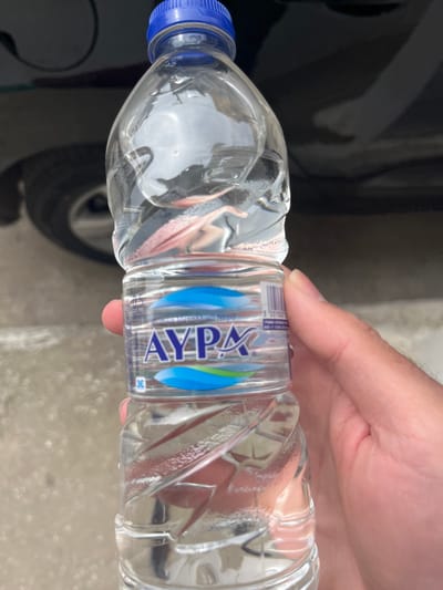 A person is holding a plastic bottle of AYPE water in their right hand. The bottle is mostly full and has a blue cap. The bottle is being held in front of a black car. The scene is set in Limenaria, Greece, at the coordinates latitude 40.627 and longitude 24.575. The pavement is gray and the car is dirty. There are no other people present.