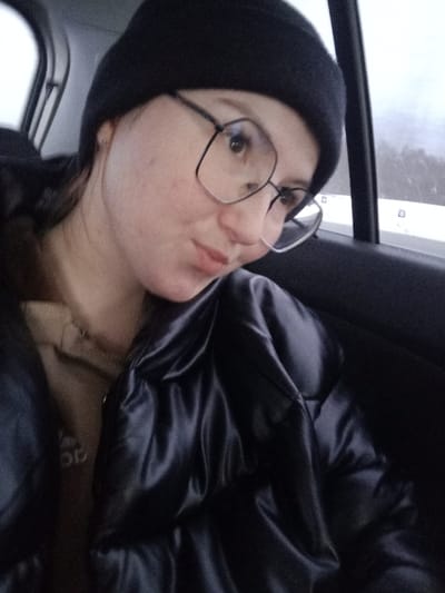 A young adult female is depicted in a close-up shot, seated within a vehicle. She wears a black knit beanie and dark-framed eyeglasses. Her fair complexion shows minor blemishes on the cheeks. A small, metallic stud earring is present in her left earlobe. She is dressed in a dark, lustrous, possibly quilted jacket, with a portion of a light brown or tan garment visible at her neck. Her expression is a subtle smirk, and her gaze is directed slightly upwards and to the right. The vehicle's dark interior is visible to the right of the subject, with a side window revealing a blurred, exterior winter landscape featuring indistinct snow-covered ground or vegetation under an overcast sky. The scene is located within a vehicle in the city of Kueda, Russia.