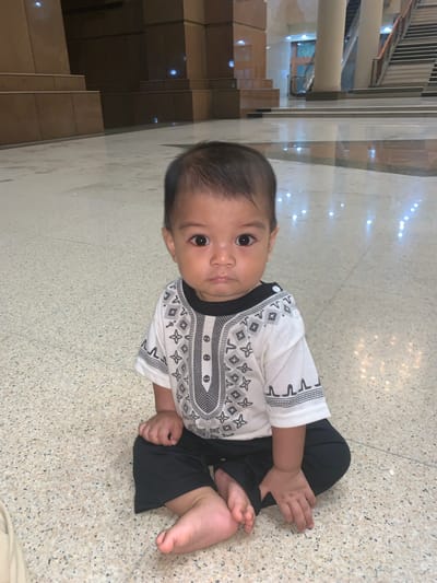 A baby is depicted sitting upright on a light-colored, speckled floor, looking directly at the camera. The baby has dark hair and dark eyes, and appears to be of Asian descent. He is wearing a white short-sleeved shirt with black and grey geometric patterns around the collar, sleeves, and down the front, paired with dark-colored pants. His hands are resting on his legs, and his bare feet are visible.

The setting appears to be indoors, likely within a building in Samarinda, Indonesia. The floor has a polished, reflective surface, showing bright, scattered light reflections. In the background, there are large, light-colored pillars and a set of stairs made of similar material, suggesting a spacious hall, lobby, or a place of worship. Further in the background, a dark wooden door or window with a patterned grate is visible. The lighting suggests it is daytime or a brightly lit interior. The baby's expression is neutral and observant. No discernible text is visible in the image.