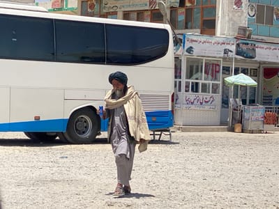 An older man wearing a white turban and a long, beige, patterned robe is walking to the left side of the image, holding a blue and white can in his left hand. His right hand is inside the folds of his robe. He is walking on a cracked, gravel surface in front of a white bus that is parked in front of a small, white building with a VIP sign.  The building has a blue and white awning over a small stand with a sign that says "Karachi Tours & Travel". The bus is facing the camera, and the front tires of the bus are visible. There are various other buildings and signs in the background. The scene is in Panjwai, Afghanistan, located at 31.618 latitude, 65.601 longitude.