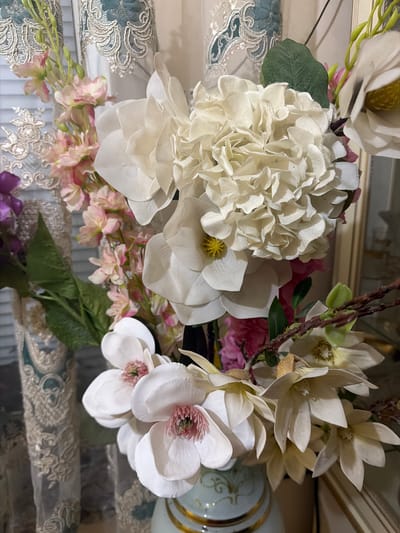 A close-up, eye-level, indoor shot focuses on an artificial floral arrangement. The arrangement features a variety of flowers, predominantly white and pale pink, with some green foliage. Notably, there's a large white hydrangea, several white magnolias with pink stamens, and smaller delicate pink blossoms. The flowers are arranged in a vase, partially visible at the bottom of the frame, which appears to be light blue with gold trim. Behind the flowers, a blurred background consists of what looks like ornate lace curtains in shades of cream and teal, and blinds. The lighting is soft, suggesting an indoor setting, possibly a home or a decorated commercial space. The image does not depict any specific location within the city of Dzhalgan, Russia, or any people.