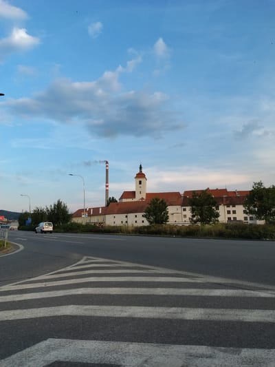 The image captures a street-level view of a city in Strakonice, Czechia, with a large complex of buildings featuring red-tiled roofs and a prominent clock tower in the background. To the left of the buildings, a tall industrial chimney emits a plume of smoke. A white van is parked on the side of the road, and a pedestrian crossing with white stripes is visible in the foreground. The sky is partly cloudy with a light blue hue. The scene appears to be during the daytime, possibly late afternoon, given the lighting.