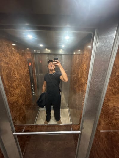The image captures a selfie taken by a young man inside an elevator. The man is reflected in a highly polished, mirror-like surface, likely the elevator door or a mirrored wall. He is dressed casually in a black t-shirt, black trousers, and a black baseball cap worn backward. With his right hand, he holds a smartphone pointed towards the reflection, his mouth slightly open in an expression for the photograph. He also wears a thin necklace and an earring.
The elevator interior showcases a mix of materials. The wall to the viewer's left (which appears as the right wall in the man's reflection) is made of brushed stainless steel. The opposite wall, where the selfie is taken, is a reflective, mirrored surface. On this mirrored surface, an elevator control panel is visible, featuring multiple buttons and a digital display. The display clearly shows the number "3," indicating the current floor. A silver handrail is also present along this wall.
The back wall and ceiling of the elevator are covered in a distinctive, dark brown, burled wood-like paneling, providing a warm contrast to the metallic elements. Artificial lighting, likely from embedded fixtures, illuminates the space and creates bright reflections on the shiny surfaces. The overall setting is a modern and clean elevator, consistent with a building in Noisy-le-Grand, France.