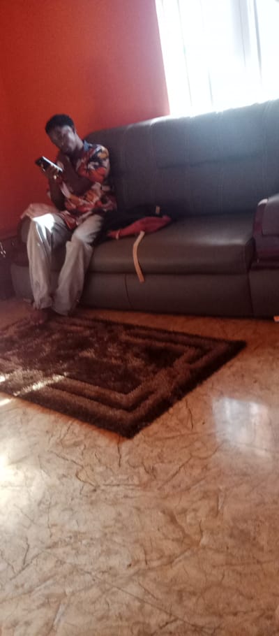 The scene depicts an interior room in Udi, Nigeria. A single individual, appearing male, with short dark hair, is seated barefoot on a dark grey multi-seater sofa. The individual wears a multi-colored patterned shirt and light-colored trousers, holding a dark-colored smartphone with both hands and looking down at the device. A black backpack with a red flap and a light-colored strap rests on the sofa to the individual's right. The wall behind the sofa is painted a solid orange-red. A bright, overexposed window or light source is visible on the right, illuminating a portion of the sofa, the individual's leg, and casting a rectangular light patch onto the floor. The floor consists of light-colored, textured tiles or marble with dark veining. A rectangular dark brown shaggy rug, featuring a raised geometric pattern, is placed on the floor in the foreground.
