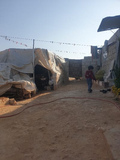 This media file captures an outdoor scene in Killi, Syria, under bright daylight with a clear, blue sky. The foreground and mid-ground consist of dusty, reddish-brown earth. A red hose or cable runs diagonally across the ground from the lower-left to the upper-right.
In the mid-ground, a young child, wearing a red long-sleeved top and dark trousers, stands with their back to the viewer, looking towards the left side of the frame. To the left, a large, makeshift structure or shelter is visible, constructed from white tarpaulin or canvas sheets, with scattered bricks and debris around its base. Above and behind this shelter, concrete block structures and a dark corrugated metal roof are partially visible.
Several lines, possibly clotheslines, are strung across the scene at various heights, adorned with numerous clothes pegs. In the background, to the right of the child, are more rudimentary structures, including what appears to be a wall and some green foliage. The scene suggests an informal settlement or a basic residential area with limited infrastructure. No specific activity beyond the child standing is evident. There is no visible text.