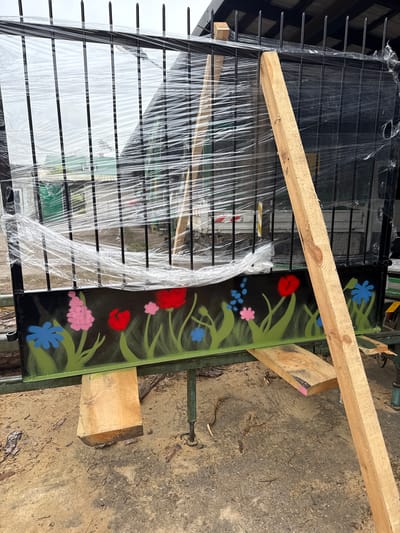 A wrought iron fence lies on its side on a dirt and concrete ground, held up by wooden blocks and planks. The fence is partially wrapped in clear plastic. One panel is painted with a black background and decorative flowers in blue, red, and pink. The setting appears to be outdoors, possibly a storage or work area. There is no clear indication of a specific location in Otwock, Poland, though the environment suggests a potentially residential or industrial neighborhood.