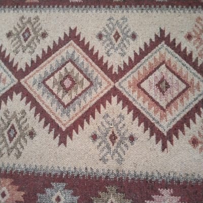 This is a close-up, overhead shot of a woven textile, likely a rug or blanket, displaying a geometric pattern. The primary colors are a neutral beige or cream as the background, accented with shades of maroon, brown, grey, and a hint of pink. The pattern features repeating diamond shapes and zig-zag or sawtooth motifs, characteristic of traditional indigenous weaving styles. The texture of the weave is clearly visible, suggesting a natural fiber material. There are no people, specific objects, or a discernible scene depicted; the focus is solely on the decorative pattern of the fabric. The lighting is even and appears to be artificial, with no indication of time of day or weather conditions. The location context of Kalachinsk, Russia, does not provide any direct visual cues within the image itself. No text is visible.
