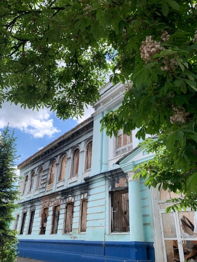 The image shows a building in Izium, Ukraine, located at the coordinates 49.192, 37.279. The building is light blue with white trim and a dark blue base. There are large windows on the side of the building, many of which are broken or boarded up. The building appears to have been damaged by fire, with soot marks on the walls. The image is taken from a low angle, looking up at the building. There are branches of a leafy tree in the foreground. The tree is in bloom and has white flowers. The sky is a light blue with some white clouds. There are no people visible in the image.